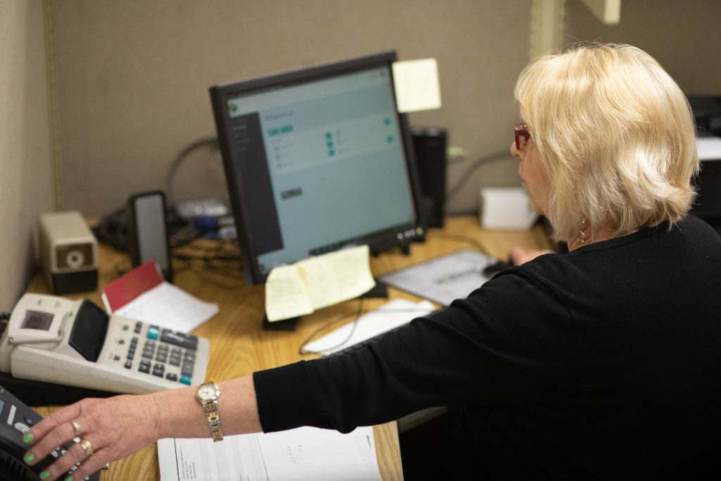 A woman reaching for the phone as she sits in an office in front of a computer.